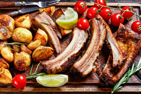 grilled pork ribs served with roast new potatoes and fresh tomatoes on a wood chopping board on a dark wooden table, horizontal view from aboveの写真素材