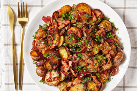 bratkartoffelsalat, roast potato salad with red onion and bacon on a white plate on a wooden table,...