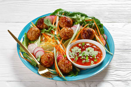 Vietnamese banh mi salad with pork meatballs, carrot, cucumber, and radish served over romaine lettuce salad on a blue plate with sweet chili sauce on white wooden background, top view, close-upの写真素材