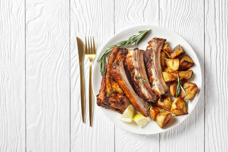 grilled pork ribs served with roast new potatoes on a white plate on a wooden table, horizontal view from above, flat layの写真素材