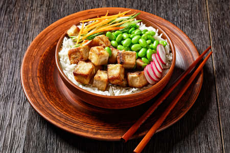 vegetarian tofu poke bowl with basmati rice, edamame beans, radish, thinly sliced cucumber and carrots salad in a clay bowl with chopsticks, horizontal view from aboveの写真素材