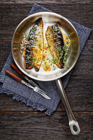 fried mackerel fillets with spices and herbs in a skillet on a dark wooden table, vertical view from aboveの写真素材