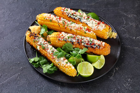 Elote on a black plate, Grilled Mexican Street Corn, charred cobs are slathered in sour cream based...