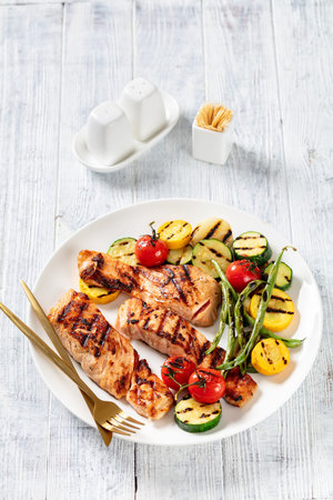 barbequed salmon pieces and grilled vegetables, zucchini, tomatoes and green beans on a white plate...