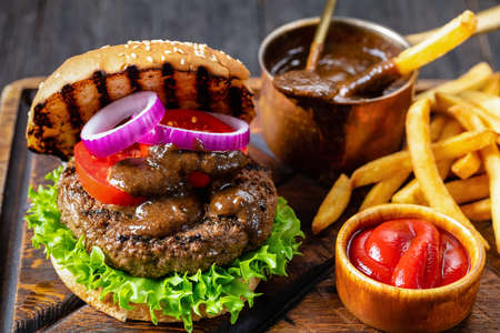 grilled hamburger of sliced bun topped with juicy beef burger, lettuce leaves, tomatoes, red onion rings and barbeque sauce topping on a cutting board with french fries and ketchup on side, close-upの写真素材