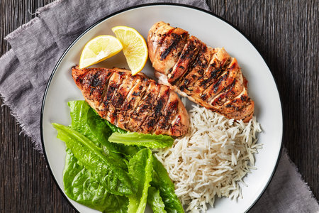 juicy grilled chicken breasts with fresh romaine lettuce leaves and rice on a plate on a dark wooden table, flat lay, close-upの写真素材