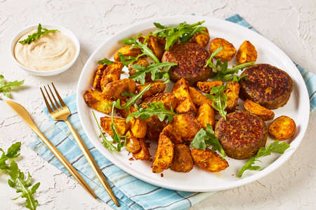 vegan lentil and bean burgers patties with baked potato wedges,  rocket leaves and hummus on a white plate on a light textured table, vegan recipeの写真素材