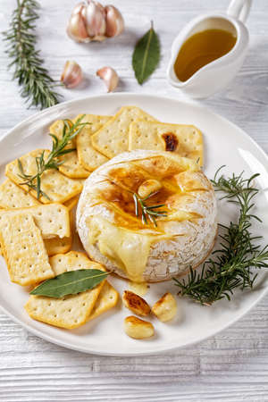Baked camembert french soft cheese topped by olive oil with rosemary sprigs, garlic cloves, and bay leafs served with crackers on a white plate on a white wooden background, top view, close-upの写真素材