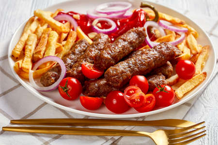 cevapcici, grilled balkan sausages with potato fries, red onion rings, red pepper, tomatoes on a white plate on a wooden table, close-upの写真素材