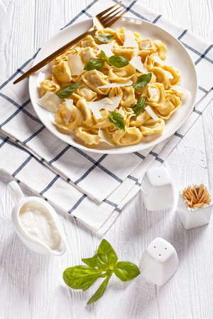 tortelloni with cheese and spinach filling, italian dumplings on a white plate with cream sauce in a gravy boat on a white wooden table, vertical view from aboveの写真素材