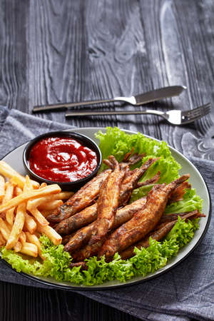 crispy fried capelin fish with fresh lettuce, french fries and tomato sauce on a plate on a black textured wooden table, vertical viewの写真素材