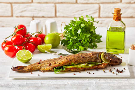 whole grilled trout fish with herbs and spices, served on a white rectangular platter with lime slices and parsley in front of brick wallの写真素材