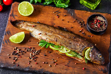whole trout fish baked in an oven with herbs and spices, served on a wooden cutting board with lime slices and parsley, close-upの写真素材