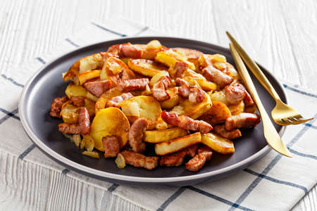 Bratkartoffeln, fried Potatoes with Bacon and onion on a plate with cutlery on a white wooden table, German cuisineの写真素材