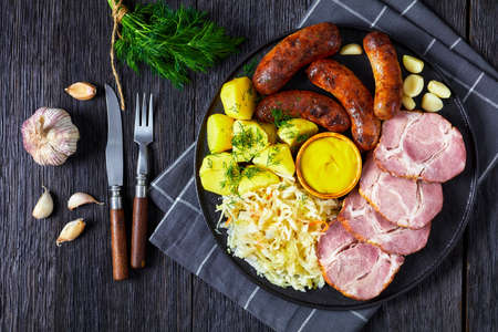 Schlachtplatte, fried bratwurst, smoked pork neck, boiled potatoes, sauerkraut and mustard on a black plate, german cuisine, horizontal view from above, flat layの写真素材