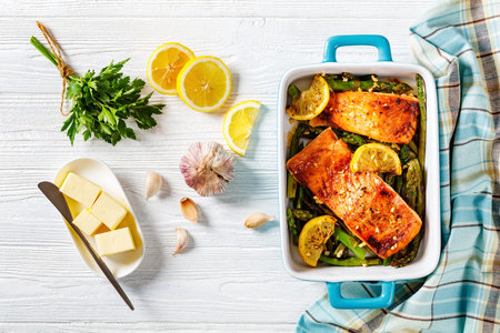 roast salmon fish fillets with asparagus and lemon in a ceramic baking dish, on white wooden table with ingredients, horizontal view from above, flat lay, free spaceの写真素材