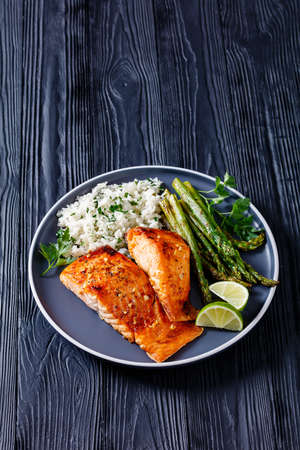 fried salmon fish fillets with asparagus, jasmine rice and lime on a plate on a black wooden table, vertical view from above, close-upの写真素材