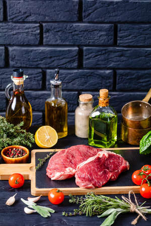raw striploin steaks on a wood board on a kitchen table with ingredients for cooking and black brick wall at the background, vertical viewの写真素材