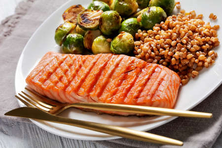 grilled salmon fillet, roast brussel sprouts and boiled farro on a white plate with golden cutlery, healthy dietの写真素材