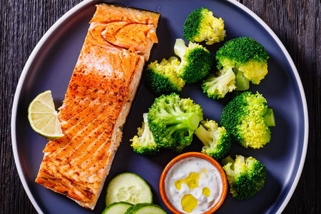 roast salmon fish fillet with boiled broccoli, fresh cucumber slices and sour cream sauce on plate, horizontal view, flat lay, close-upの写真素材