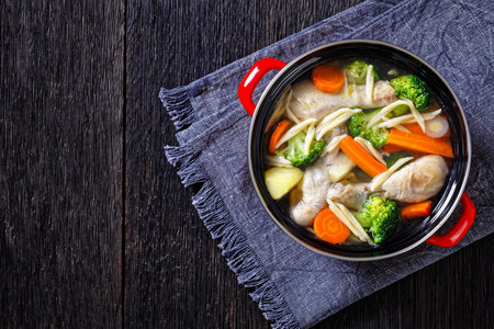 chicken spring soup with broccoli, carrots, parsnip, leek and pasta in red pot on dark wooden table,...