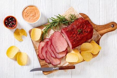 fresh sliced beef pastrami on wooden board with fresh rosemary, and potato chips on white table with...