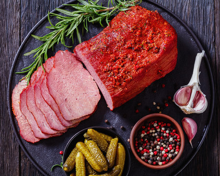 fresh sliced beef pastrami sliced on black plate with fresh rosemary, garlic and pickles on dark...