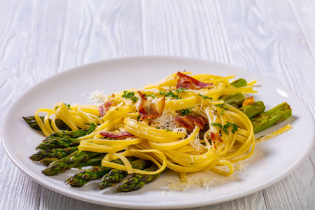 roasted asparagus spears with pasta sprinkled with shredded pecorino cheese, fried bacon pieces and parsley on white plate on white textured wood table, close-upの写真素材