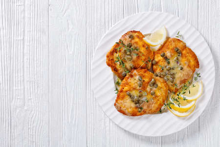 delicious chicken piccata of parmesan breaded seared chicken breasts with lemon butter caper sauce on white plate on white wooden table, flat lay, free spaceの写真素材