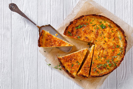 bacon and cheddar cheese quiche with hash brown crust on wood cutting board on white wooden table,...