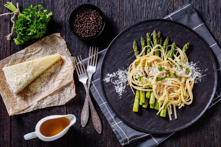 roasted asparagus spears with spaghetti sprinkled with shredded pecorino cheese and parsley on black plate on dark wooden table with ingredients, horizontal view from above, flat layの写真素材