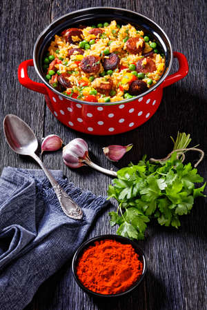 smoked sausages with rice, bell pepper, onion and green peas in red pot on wooden table, vertical viewの写真素材