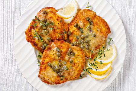 delicious chicken piccata of parmesan breaded seared chicken breasts with lemon butter caper sauce on white plate on white wood table, flat lay, macroの写真素材
