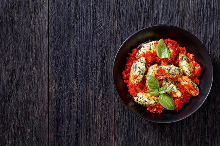 Malfatti, Italian spinach ricotta dumplings in tomato sauce with herbs and grated parmesan cheese in black bowl on dark wooden table, flat lay, free spaceの写真素材