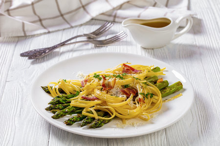roasted asparagus spears with spaghetti sprinkled with shredded pecorino cheese, fried bacon pieces and parsley on white plate on white textured wood table, close-upの写真素材