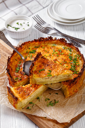 bacon and cheddar cheese quiche with hash brown crust on wood cutting board with utensil on white wooden table, vertical view from aboveの写真素材