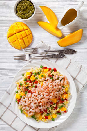 salad pink shrimps topped over millet porridge with green peas, mango, red peppers on plate on white table with pesto sauce and fresh mango, vertical view from aboveの写真素材