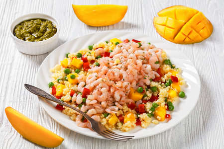 cooked salad shrimps topped on millet porridge with green peas, mango, red peppers on white plate on white wood table, with ingredientsの写真素材