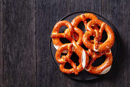 soft pretzels baked in the form of knot and sprinkled with salt on black plate on dark wooden table, horizontal view from above, flat lay, free spaceの写真素材