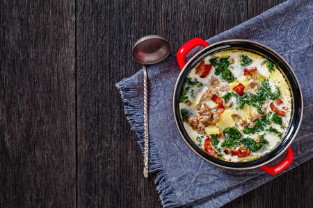 Zuppa Toscana, hearty Tuscan Soup loaded with Italian sausage, kale, bacon and potatoes in red pot with soup ladle on dark wooden table, horizontal view from above, flat lay, free spaceの写真素材