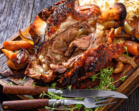 Crispy German Pork Knuckle, roast ham hock, Schweinshaxe, with Beer Gravy, Sauerkraut and roast vegetables on rude wooden serving board on dark wooden table, close-upの写真素材