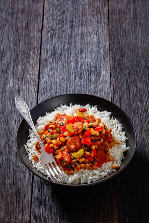 Hoppin John of black-eyed peas, bacon, sausages, vegetables and scallions with rice in bowl with fork on dark wooden table, traditional Southern american New Year dish, vertical view, free spaceの写真素材