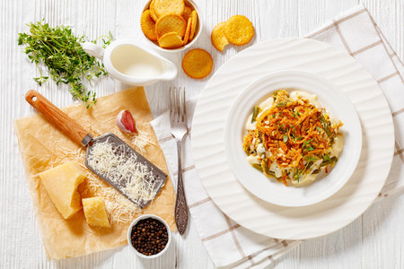 cheesy green bean casserole with fried french onion, parmesan and crackers topping on white plate on wood table with ingredients, horizontal view from above, flat lay, close-upの写真素材