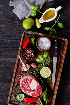 raw osso buco, veal shank steak on wood rustic cutting board with peppercorns, fresh thyme, chili peppers, olive oil, knife and lime on dark wood table, vertical view from aboveの写真素材