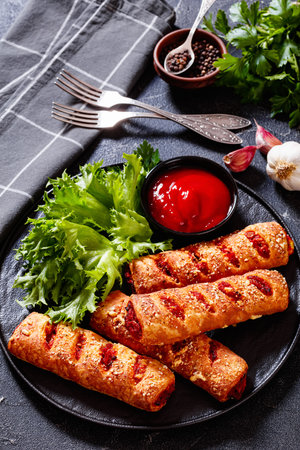 chorizo and semi-dried tomato puff pastry rolls on black plate with ketchup and fresh lettuce leaves, vertical view, close-upの写真素材