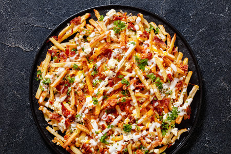 Bacon Mozzarella Feta Cheddar Cheese Fries with ranch dressing and parsley on black dish on concrete...