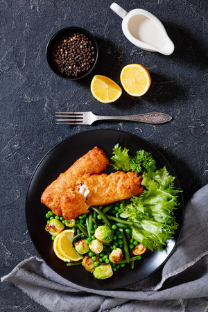 Crispy Beer Batter Cod Fish fillet with roast brussel sprouts, green beans, green peas and fresh lettuce on black plate on concrete table, vertical view from aboveの写真素材