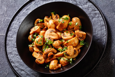 close-up of Ajillo Mushrooms, Spanish Garlic Mushrooms fried with sherry, smoked paprika and chopped parsley served in black bowl on concrete tableの写真素材