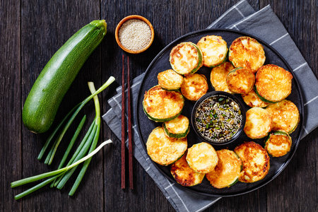 Hobak Jeon, pan fried zucchini in egg batter served with soy dipping sauce on black plate on wooden table with ingredients, korean cuisine, flat layの写真素材