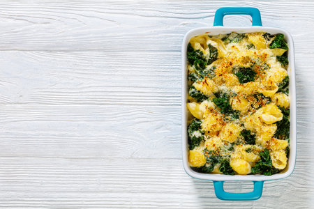 cheesy baked shell pasta with green leafy kale topped with crispy breadcrumbs in baking dish on white wood table, horizontal view, flat lay, free spaceの写真素材